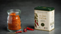 Glass jar filled with bright red chili flakes beside a beige tin labeled 'Pimentón de la Vera' with a green lid on a dark background.