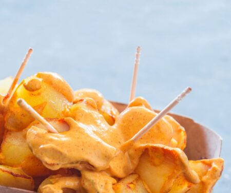 Golden potato wedges coated in creamy orange sauce with white skewers resting on a cardboard tray by a blue background